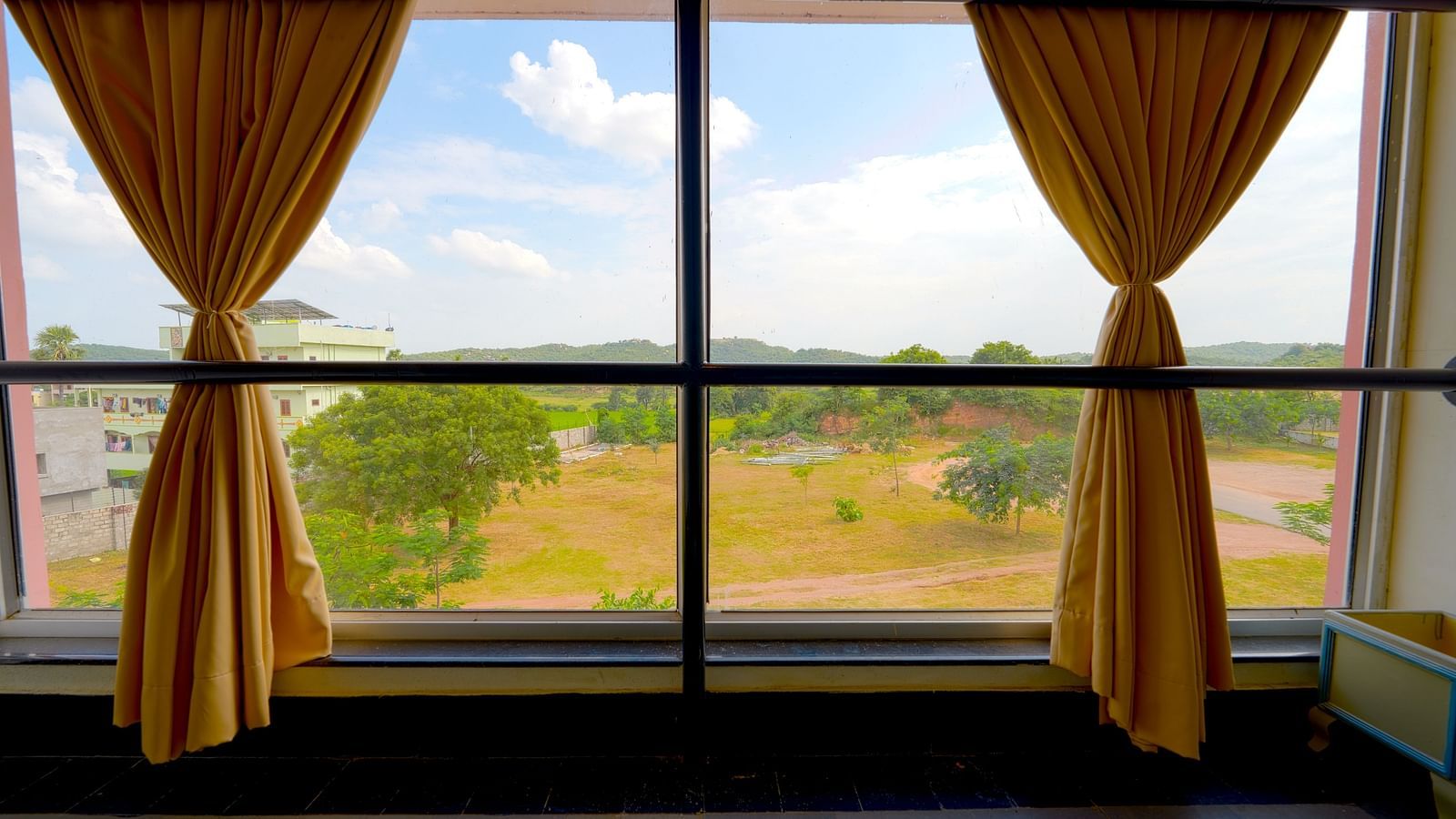 A scenic view from a hotel room window with yellow drapes overlooking a green landscape at Greens Inn - Scenic Views in Ramoji Film City.