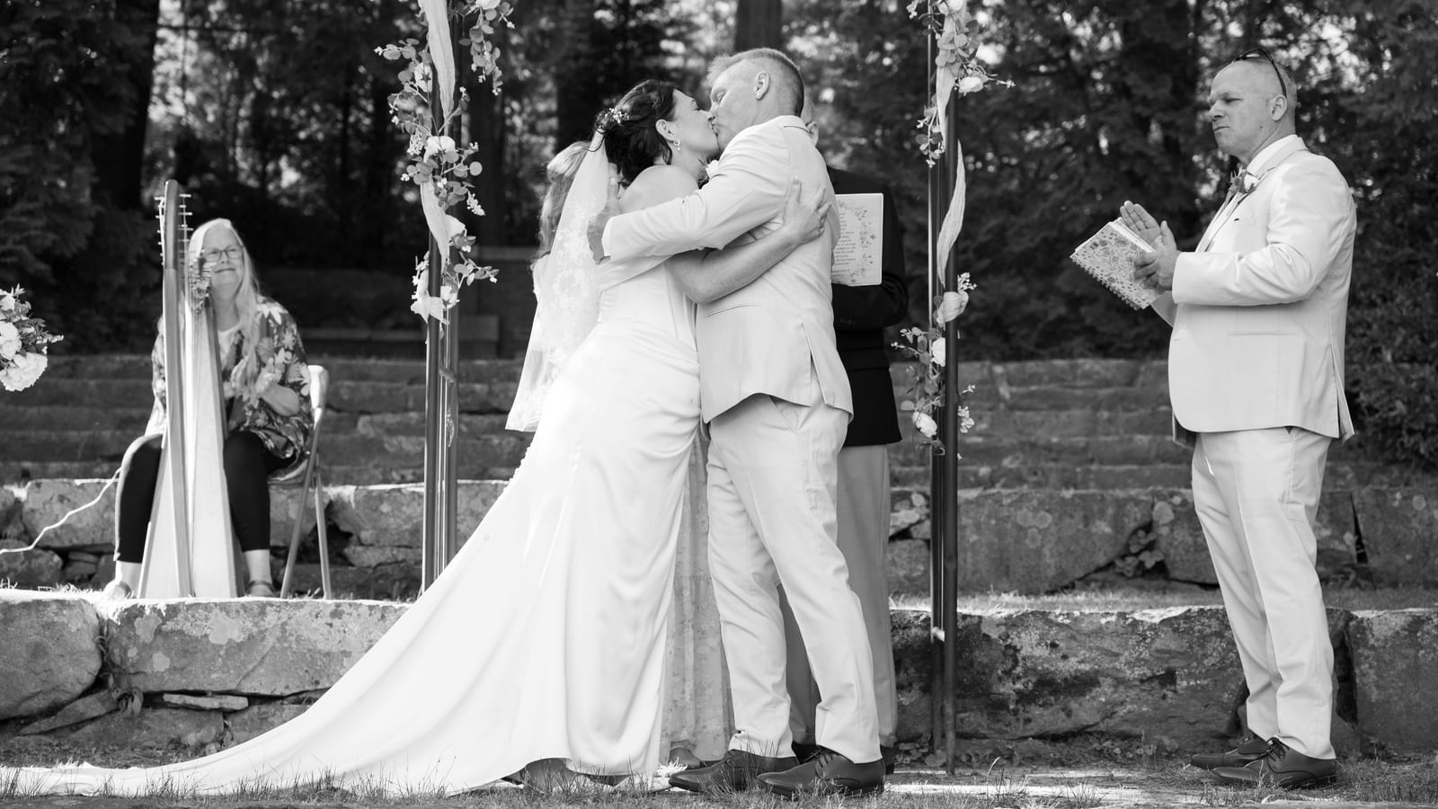 At Hartstone Inn, a couple kisses under a floral arch during an outdoor ceremony, with a harpist seated on the left and two people holding books on the right.