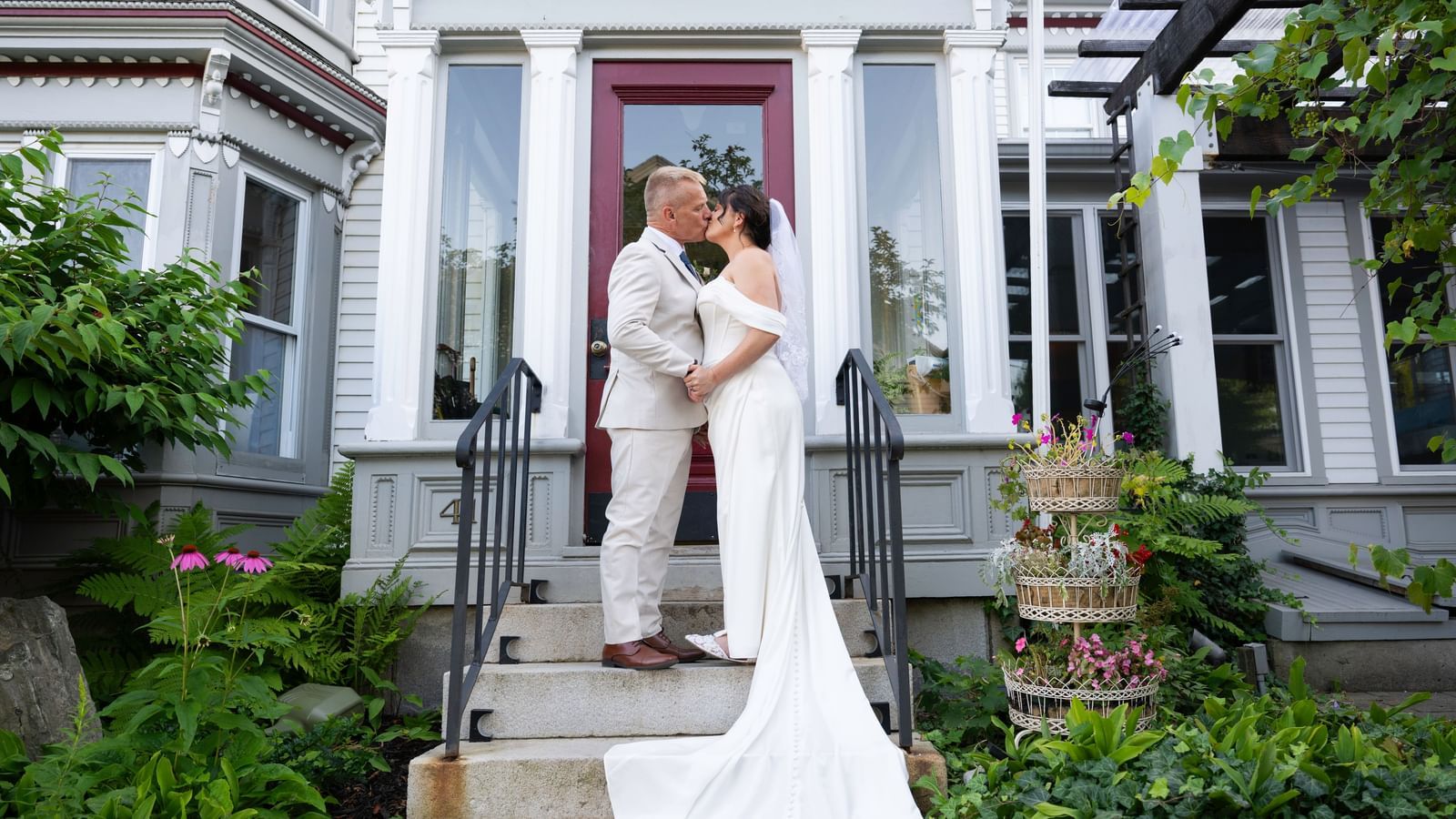 Bride and groom kissing on the front steps of Hartstone Inn, surrounded by plants and flowers, with the entrance door and windows of the building in the background.