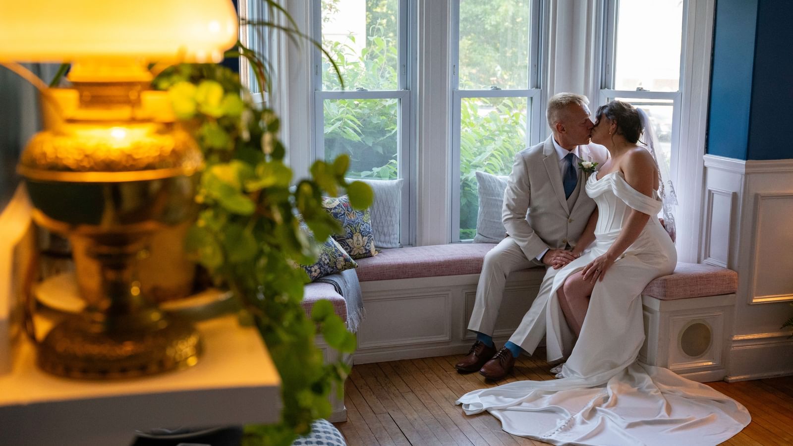 Couple in wedding attire sitting on a window seat and kissing inside Hartstone Inn, with a lit lamp, green plant, and pillows visible in the room.