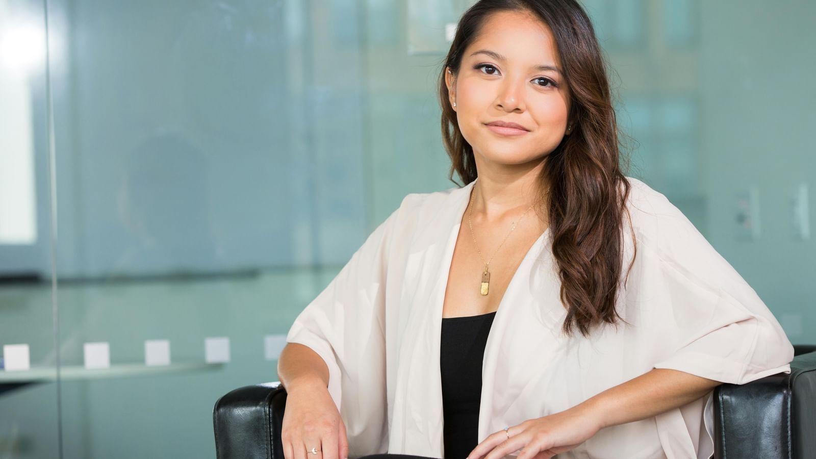 A young woman with long dark hair, wearing a light-colored cardigan over a dark top, sitting in a chair and smiling.