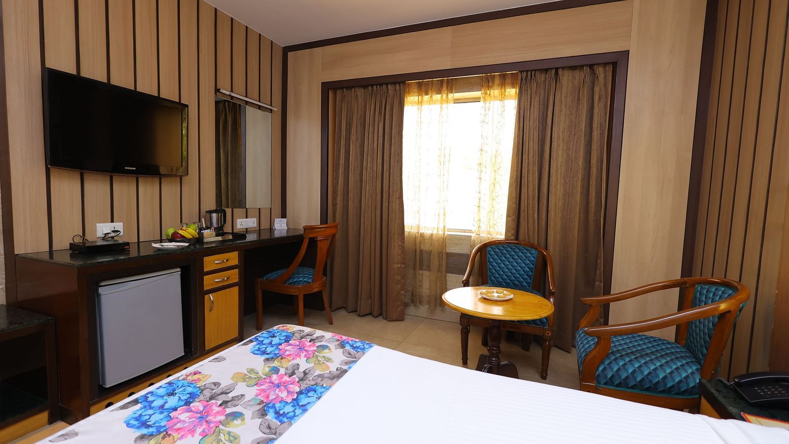 Side view image of a deluxe room at Hotel Amar with a bed, table, TV and huge window with curtains. 