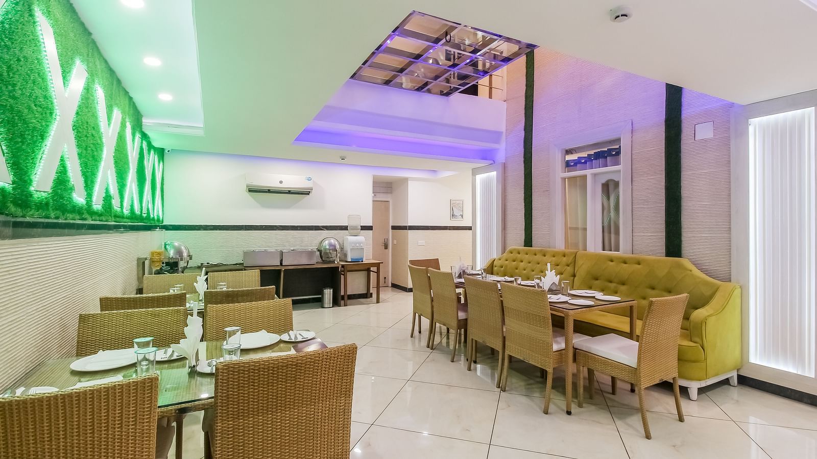 A restaurant interior with a long tufted sofa, wooden tables and chairs, a decorative wall design, and a tiled floor, lit by soft lighting. - Hotel Diamond Plaza, Chandigarh.