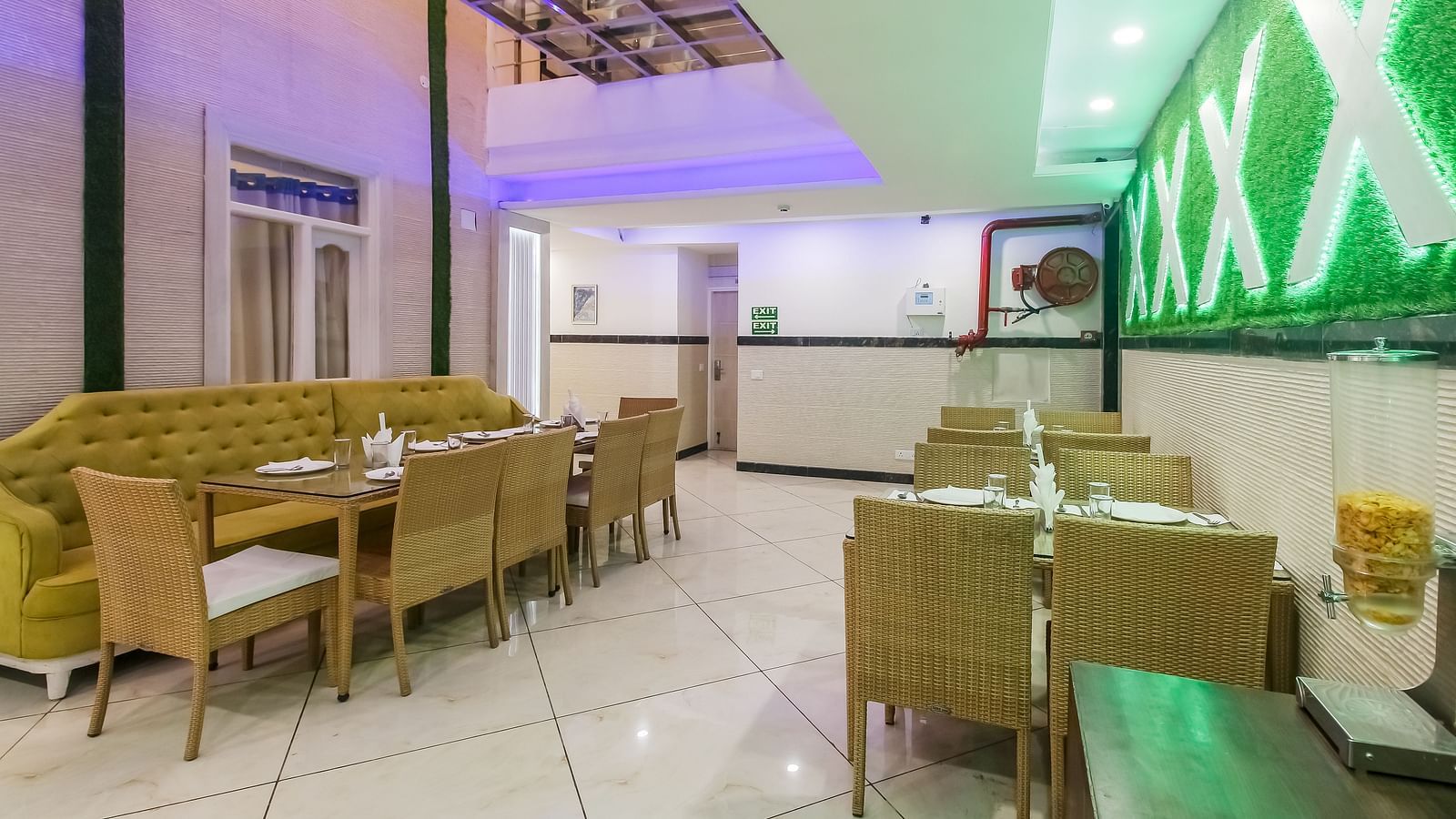 A restaurant interior with a long tufted sofa, wooden tables and chairs, a decorative wall design, and a tiled floor, lit by soft lighting. - Hotel Diamond Plaza, Chandigarh.
