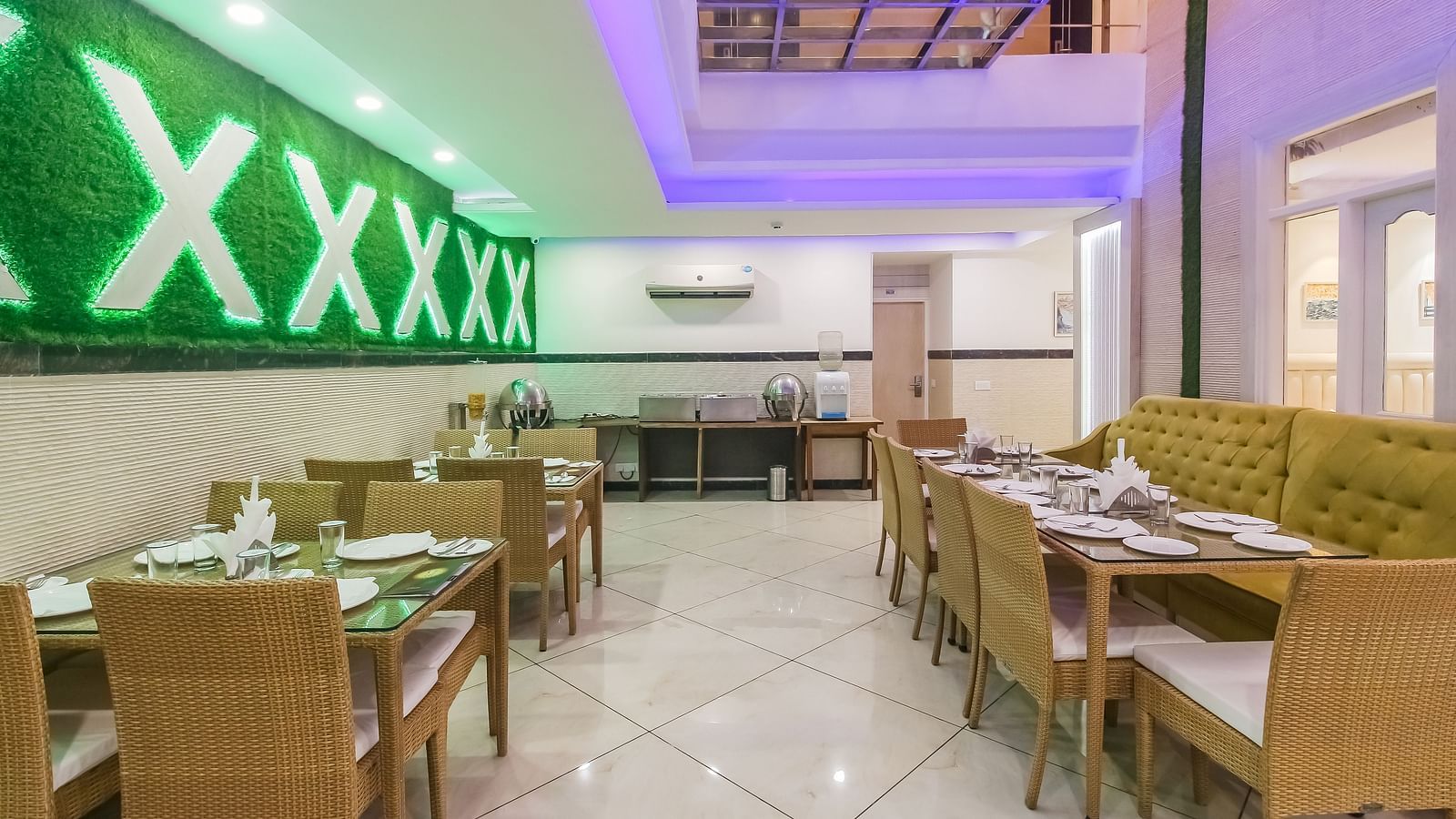 A restaurant interior with a long tufted sofa, wooden tables and chairs, a decorative wall design, and a tiled floor, lit by soft lighting. - Hotel Diamond Plaza, Chandigarh.