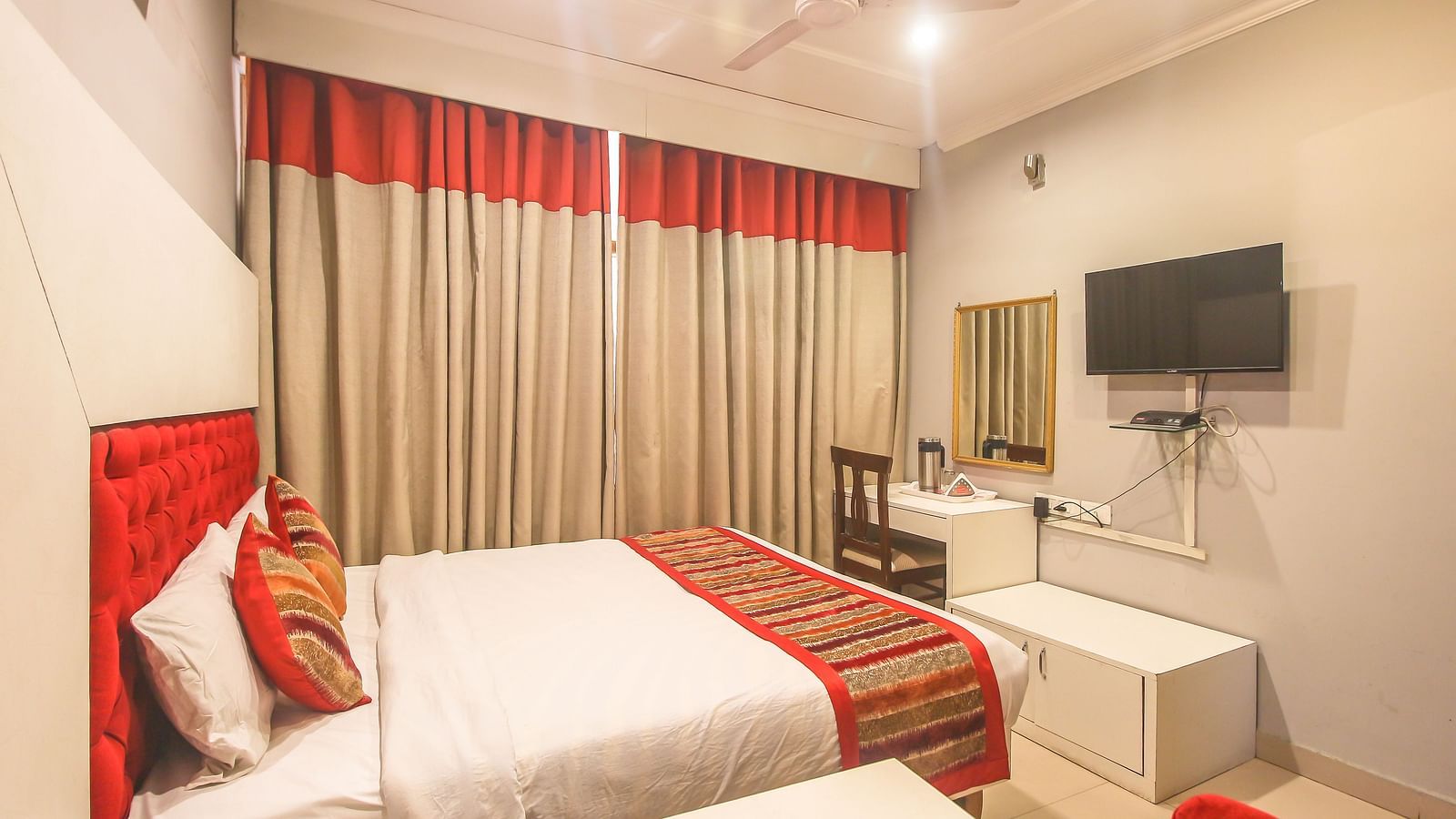 A hotel room with a plush bed, layered curtains, a TV, a desk, and a bright red accent chair, illuminated by soft ceiling lights. - Hotel Diamond Plaza, Chandigarh.