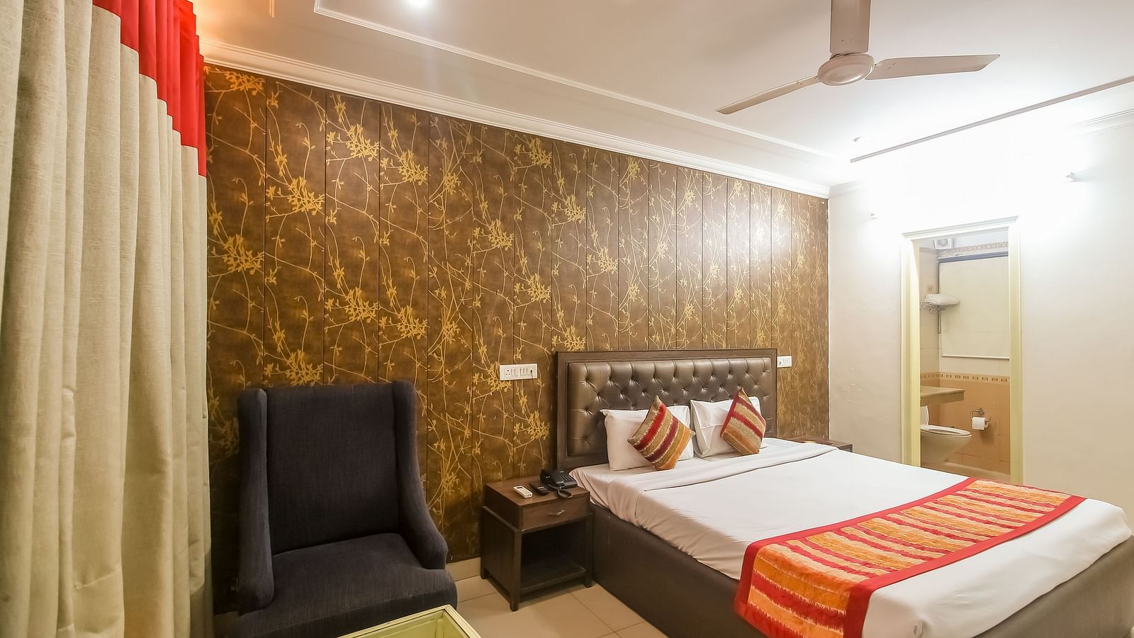 A hotel room with a textured accent wall, a plush bed with decorative pillows, a dark armchair, and a ceiling fan, illuminated by warm lighting. - Hotel Diamond Plaza, Chandigarh.