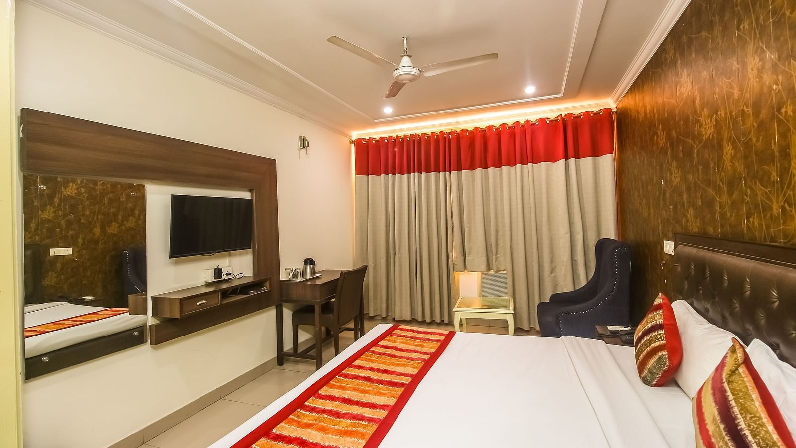 A hotel room with a plush bed featuring a patterned runner, a TV, a desk, a dark armchair, and a textured accent wall, lit by soft lighting. - Hotel Diamond Plaza, Chandigarh.