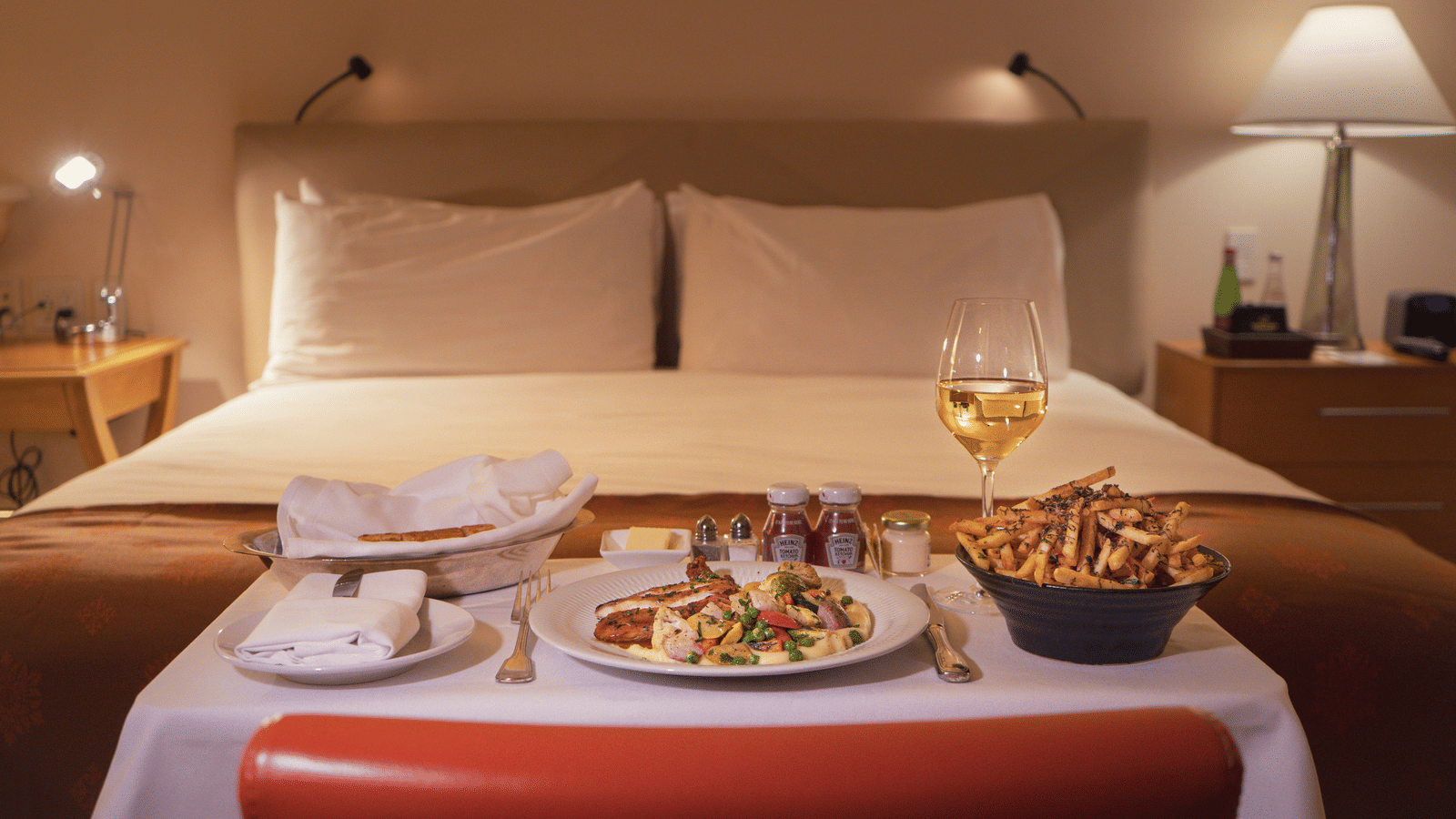 In room dining setup with a meal, fries, and a glass of wine placed on a table near a hotel bed.