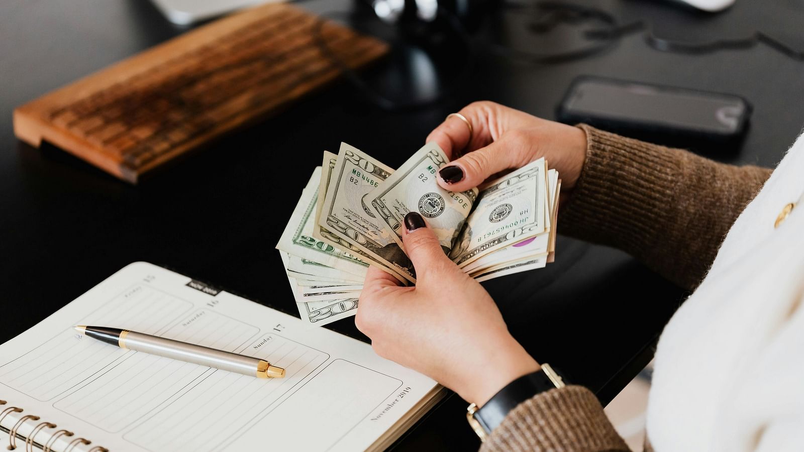 a person counting dollar notes with a notebook and pen kept on the desk with some text written on the notebook