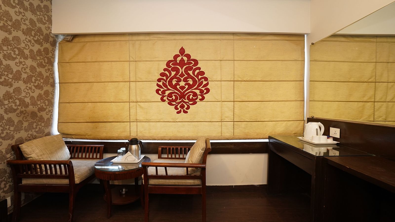 Deluxe Room featuring a seating area with chairs and a small table, a window covered by a decorative blind with a red pattern, and a desk with a kettle, set against a backdrop of textured walls and warm lighting.- Hotel Dreamland, Chandigarh