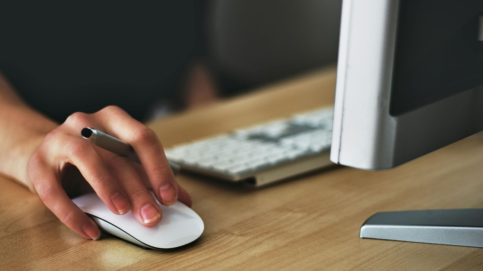 A person is using a white computer mouse on a wooden desk next to a keyboard and a computer monitor.