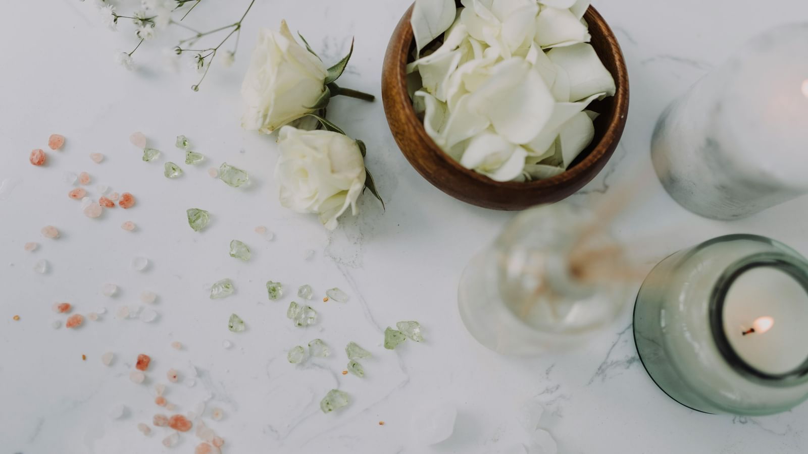 A flat-lay of spa essentials including white rose petals in a wooden bowl, white roses, bath salts, and lit candles, creating a relaxing ambience.