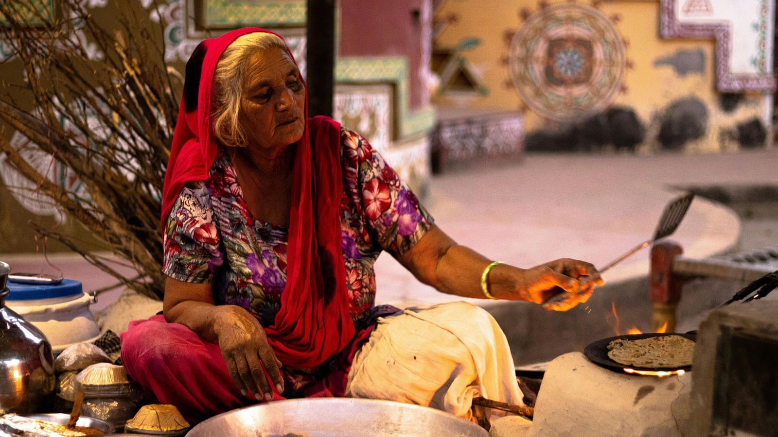 A woman in traditional attire cooks over a fire on a stove, surrounded by cooking utensils and a vibrantly decorated background.