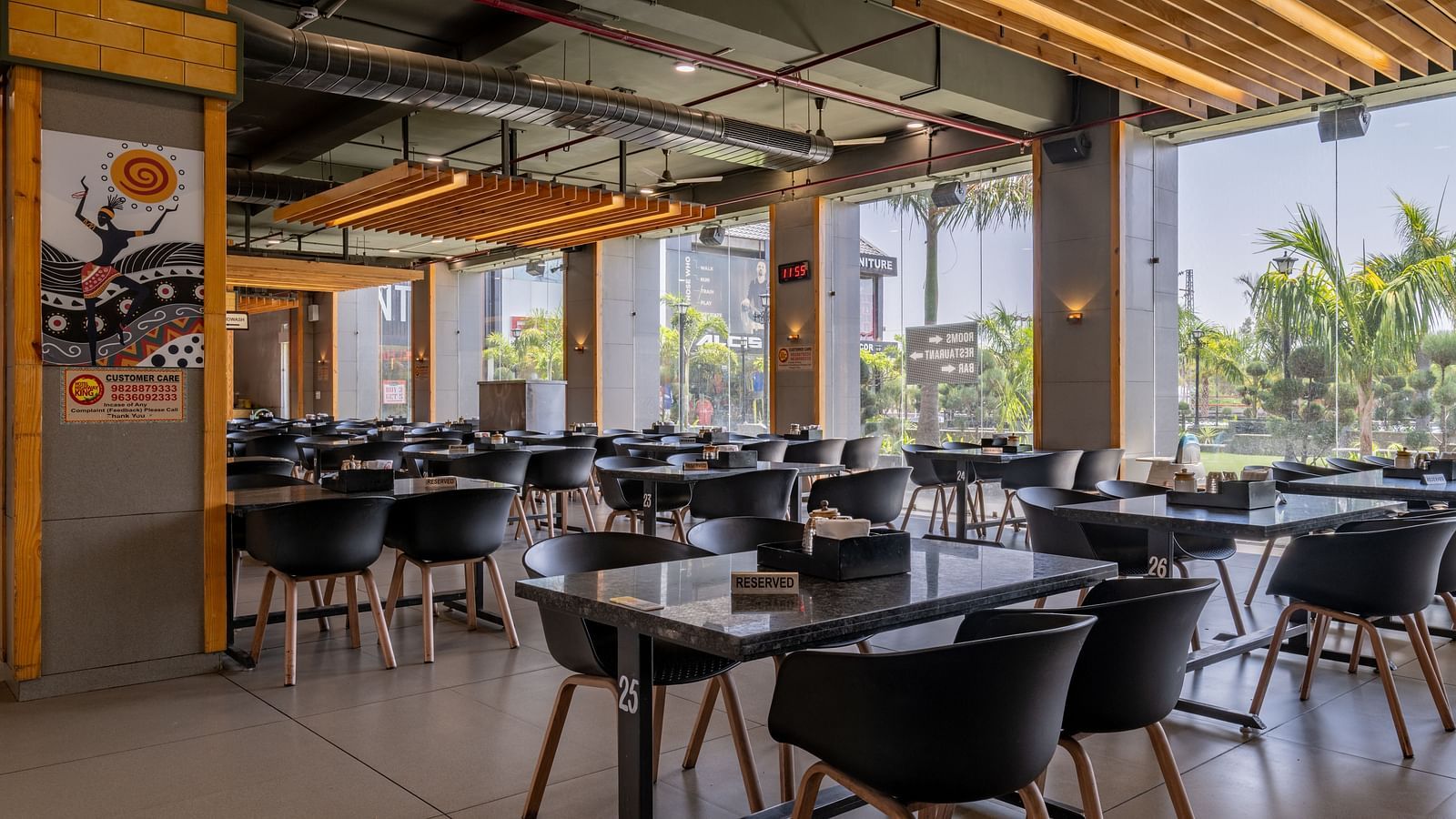 Dining area at Hotel Highway King, Bagru, with multiple tables and chairs, large windows, and wooden ceiling accents.