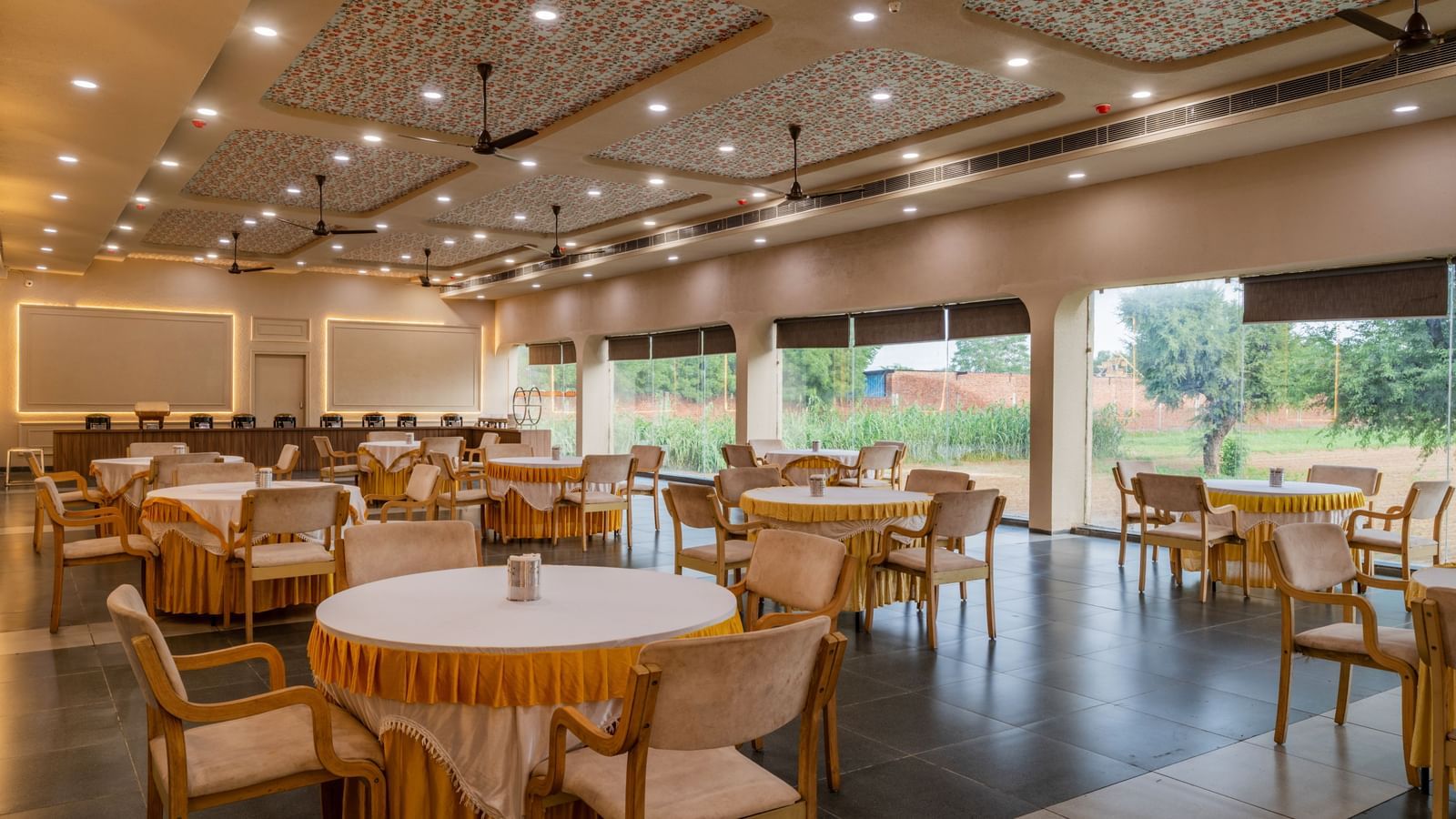 Spacious banquet area with large windows and round tables surrounded by chairs at Hotel Highway King, Neemrana. 