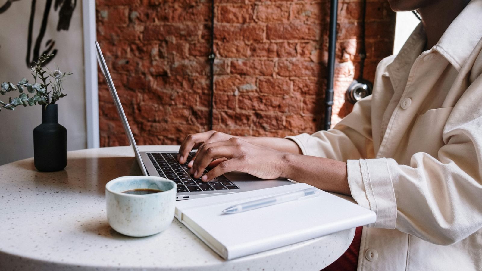 Person typing on a laptop at a round table with a notebook, pen, and a cup of coffee, against a brick wall background.