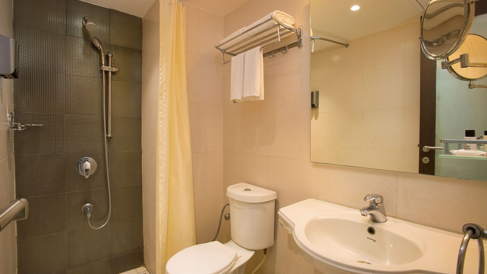 A hotel bathroom with a beige tiled shower area, white toilet, sink and a mirror, with a yellow shower curtain - Hotel Langford Keys by TGI, Shanti Nagar