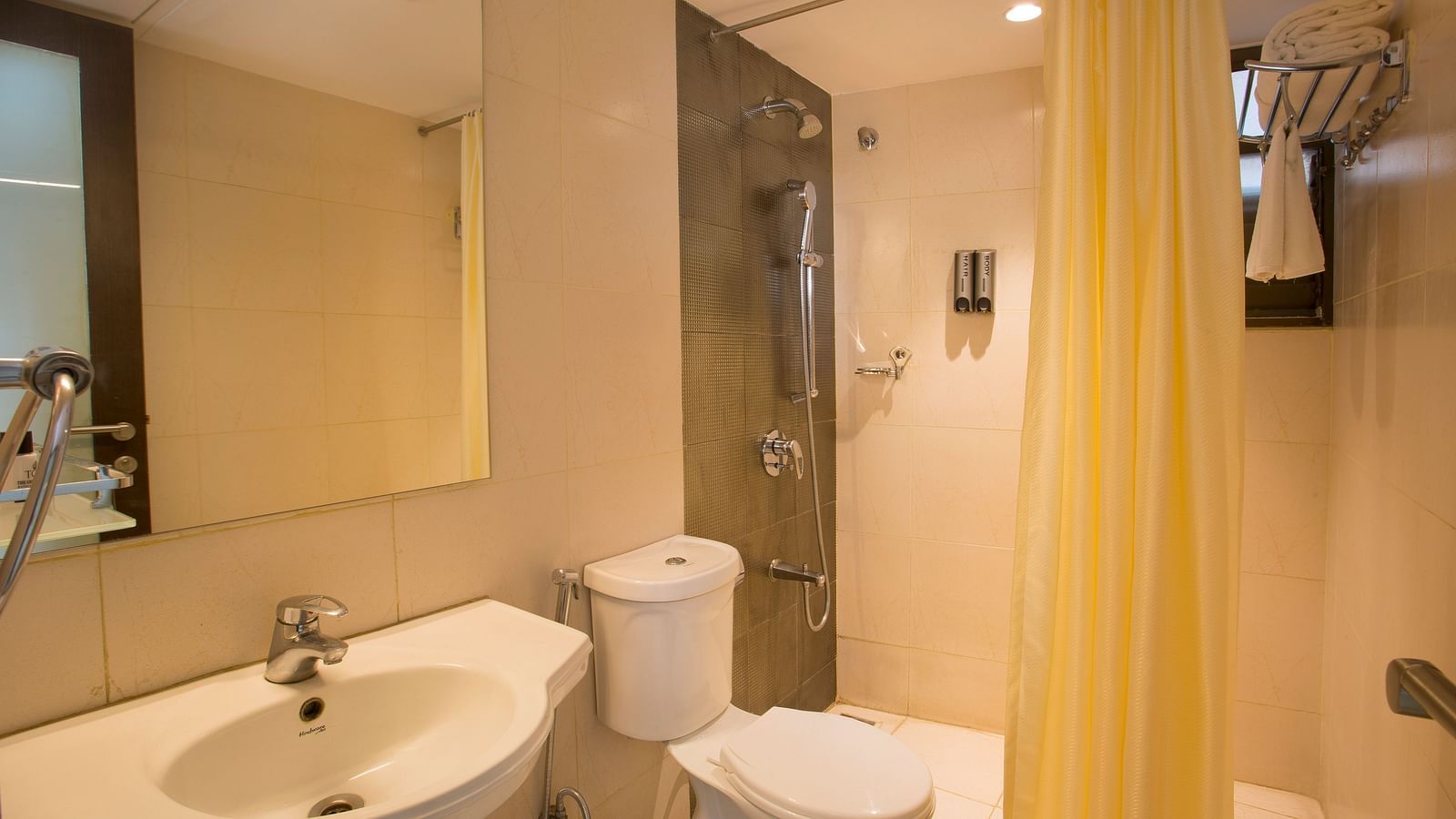 Another view of a hotel bathroom with a beige tiled shower area, white toilet, sink and a mirror, with a yellow shower curtain - Hotel Langford Keys by TGI, Shanti Nagar