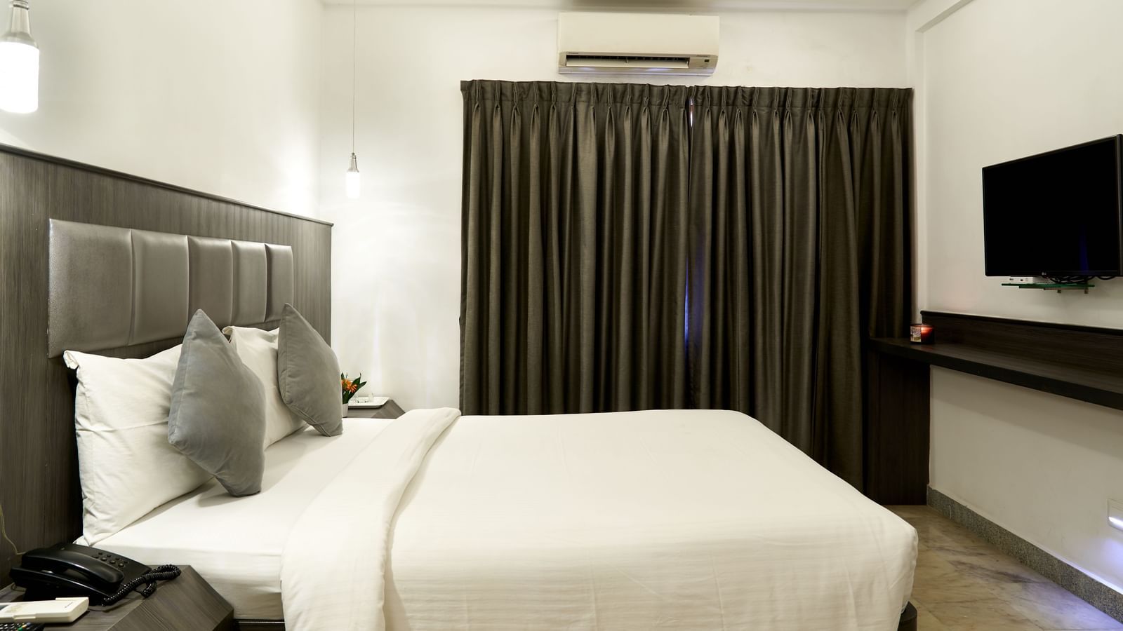 A modern hotel room at Hotel Mamallaa Heritage, featuring a bed with a dark upholstered headboard, white linens, and a flat-screen TV.