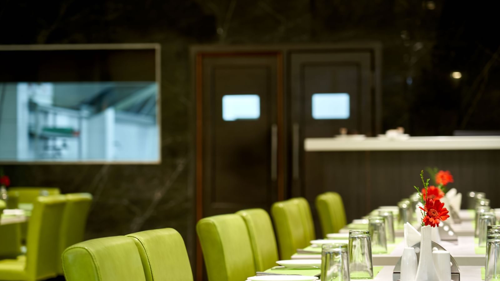 A modern dining area in Hotel Mamallaa Heritage, with a long table set for multiple guests, surrounded by lime green chairs, against a dark backdrop.