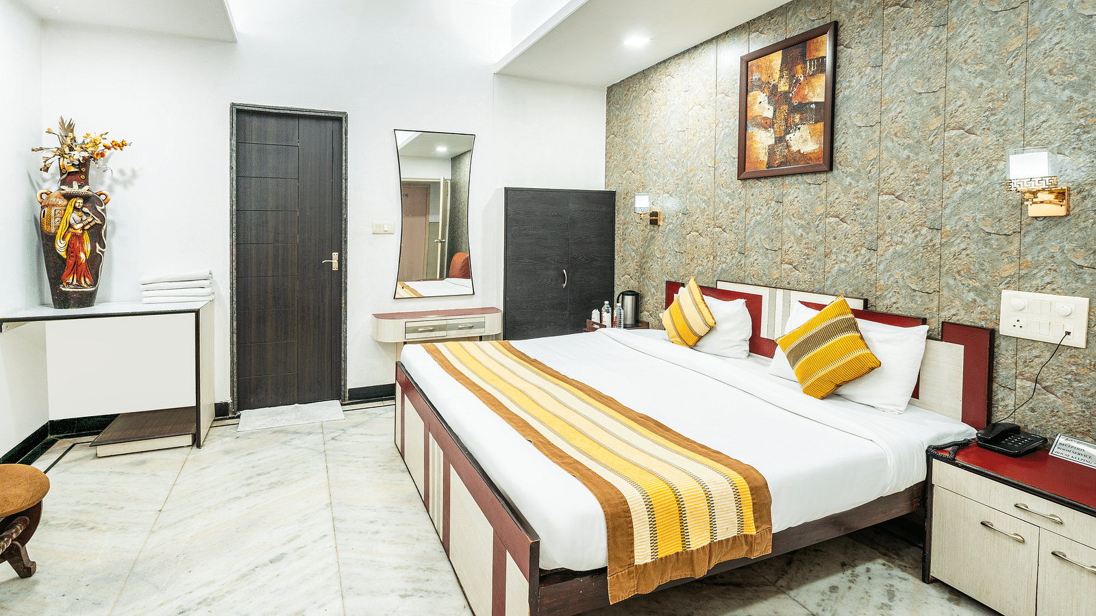 A side angle view of a room at Hotel Moti Palace, featuring a comfy bed, a painting on the wall, a flower pot, and a cupboard, creating a warm and inviting atmosphere.