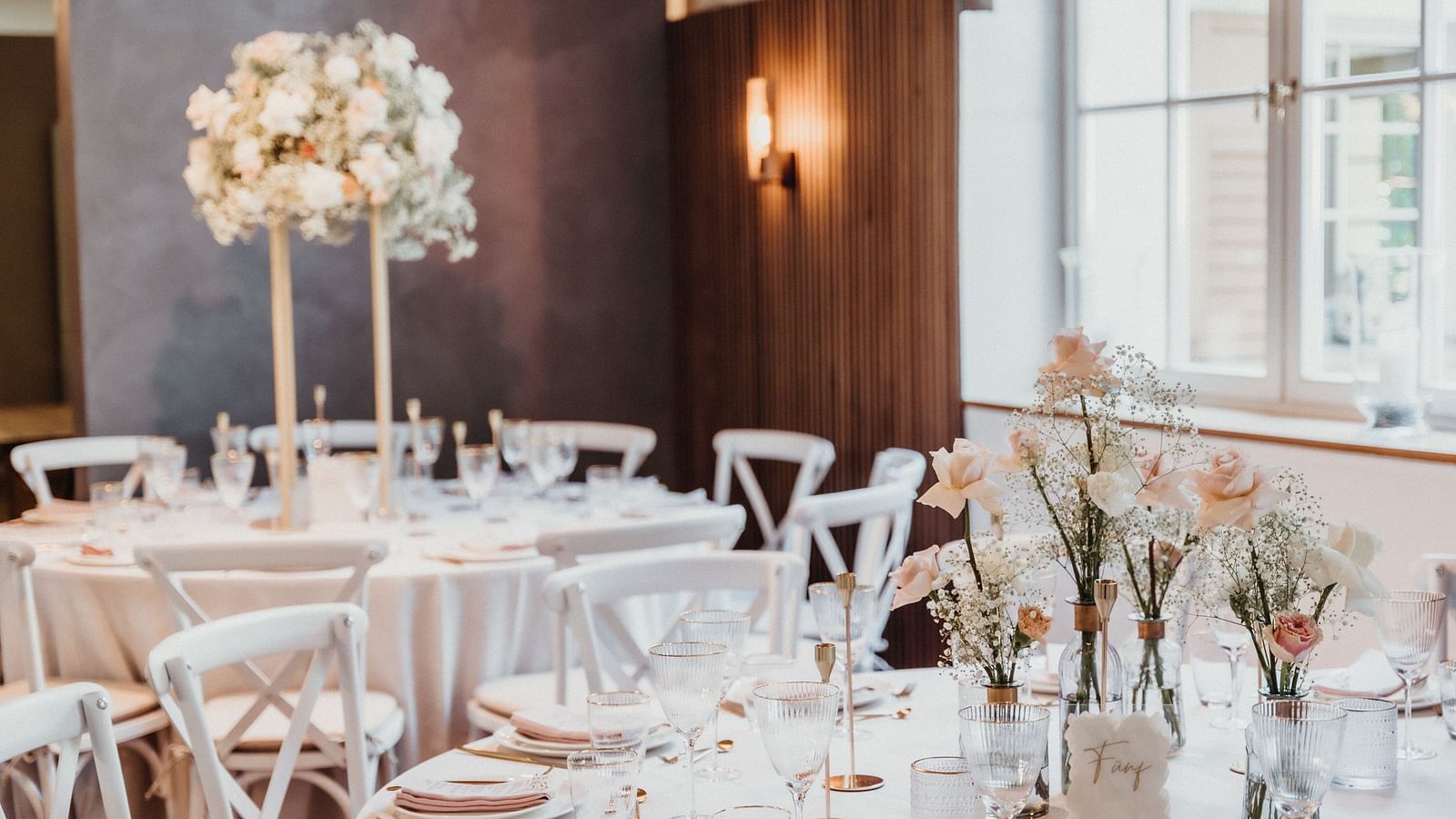 Close-up of a dining table with white chairs, floral centrepieces and delicate table settings against a dark feature wall
