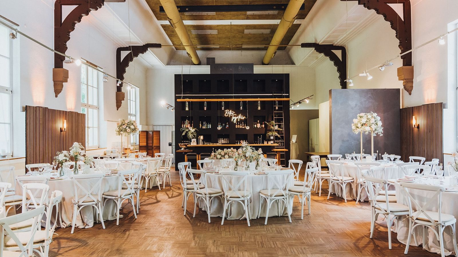 Elegantly set dining hall with white chairs, round tables with white linens and a central bar area under rustic industrial beams