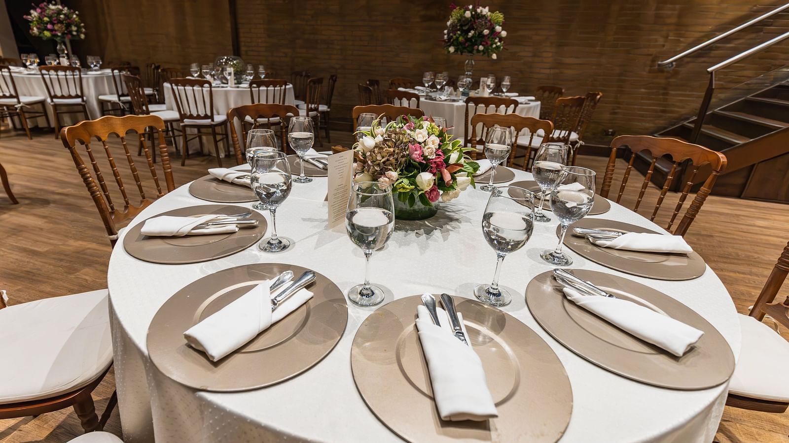 A modern dining setup with round tables covered in white tablecloths, wooden chairs and small floral centre pieces in an elegant room with a wooden wall