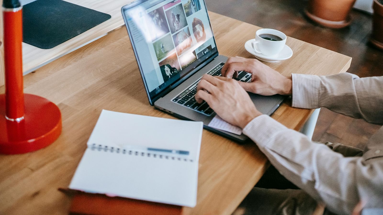 A man working on a laptop placed on a wooden table next to a coffee cup and a notebook.