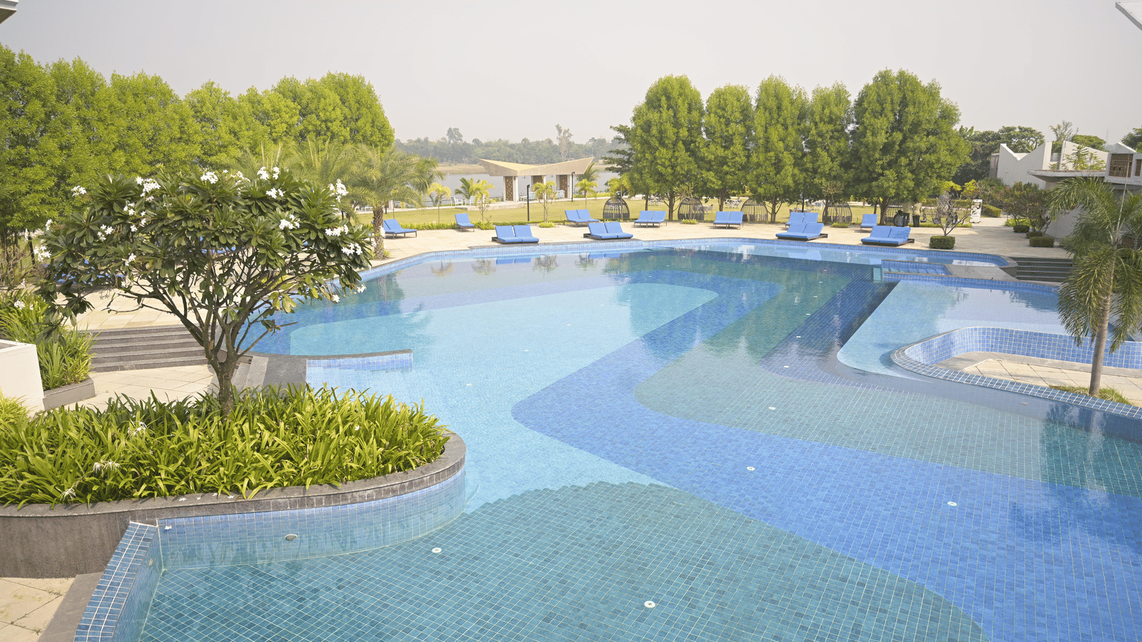 A curved swimming pool surrounded by manicured gardens and open spaces at Hotel Sonar Bangla Mayapur.