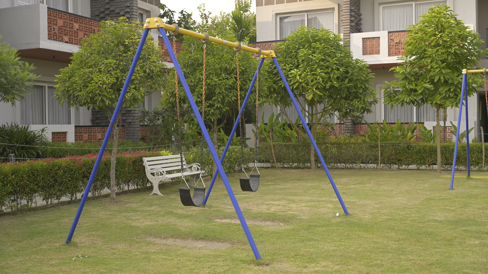 A set of blue swings placed on a green lawn surrounded by trees at Hotel Sonar Bangla Mayapur.