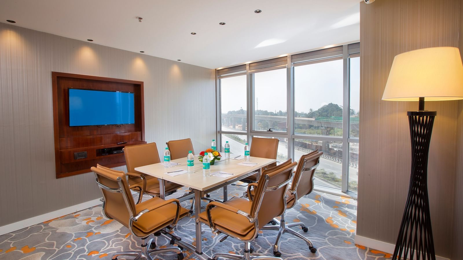 A small meeting room with a round table, tan leather chairs and a TV, with a large window and a floor lamp - Hotel Southend by TGI - Bommasandra, Bangalore