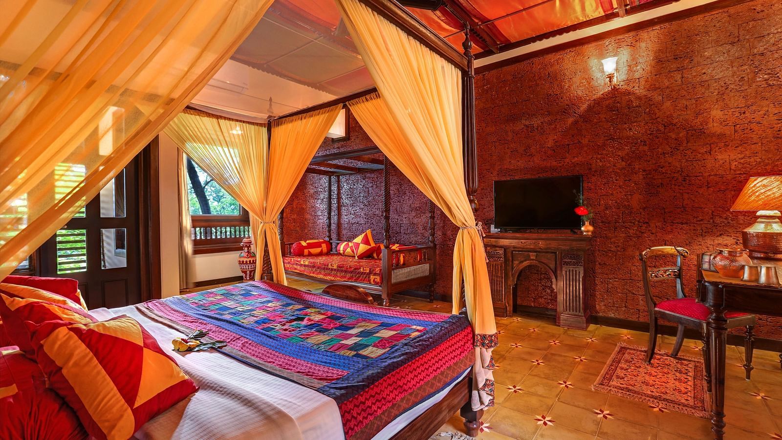 An image of a master suite bedroom with poster bed, windows, flat TV screen, wooden table, lamp, and many more - Hoysala Village Resort, Hassan