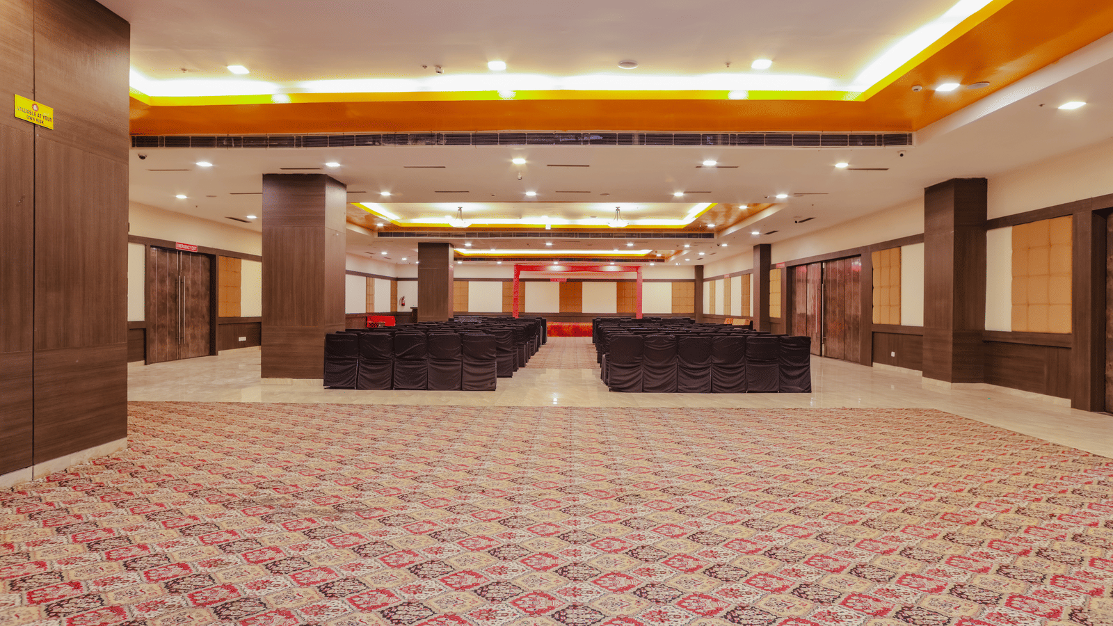 A large, empty banquet hall or conference room with patterned red carpet, a high ceiling with recessed lighting, and a stage or raised area at the far end.