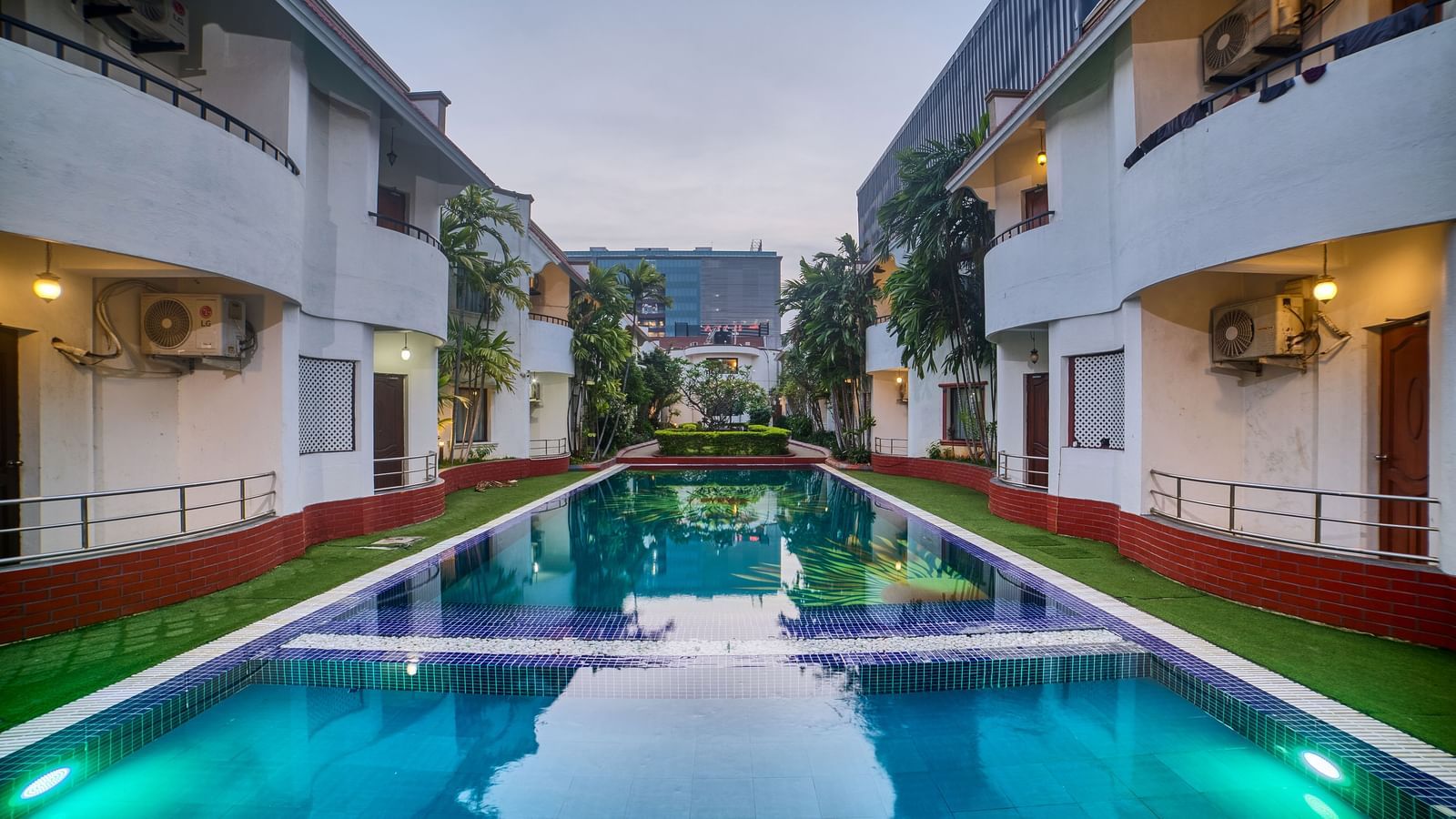 A symmetrical view of Ilara Hotels & Spa, OMR, Chennai illuminated at dusk, lining a long, reflective swimming pool or water feature.