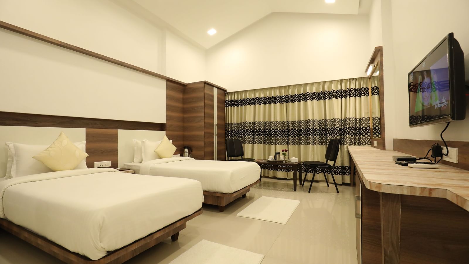 A room in Imperial Grande with twin beds, featuring a wall mounted television, and a large window with its drapes closed.