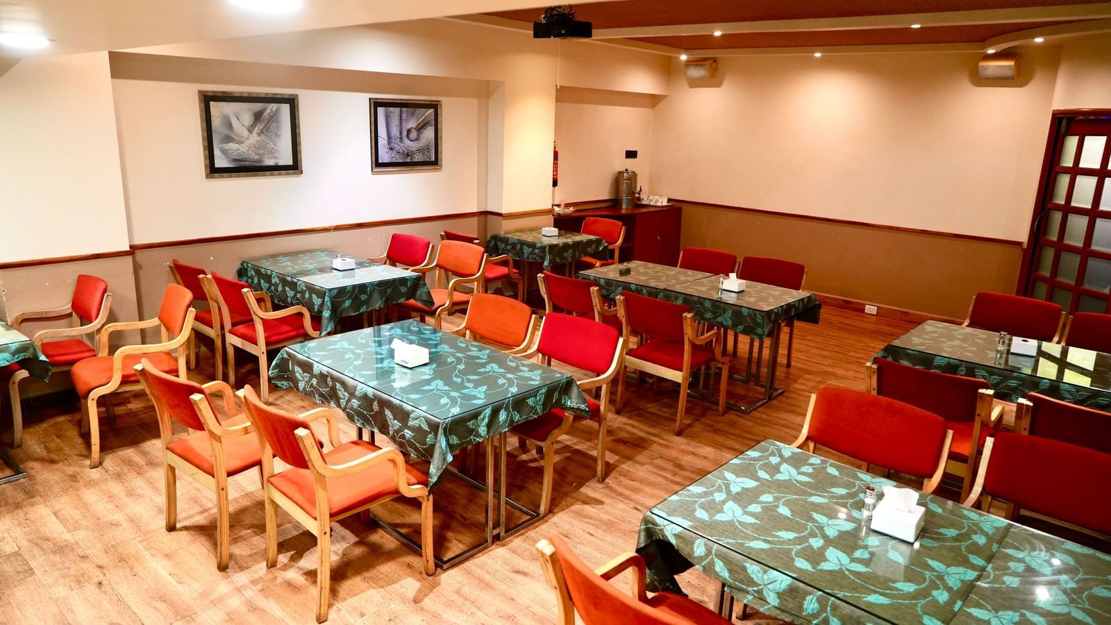 An empty dining room with multiple tables, patterned tablecloths, and several upholstered chairs at Infiniti Hotel, Indore.