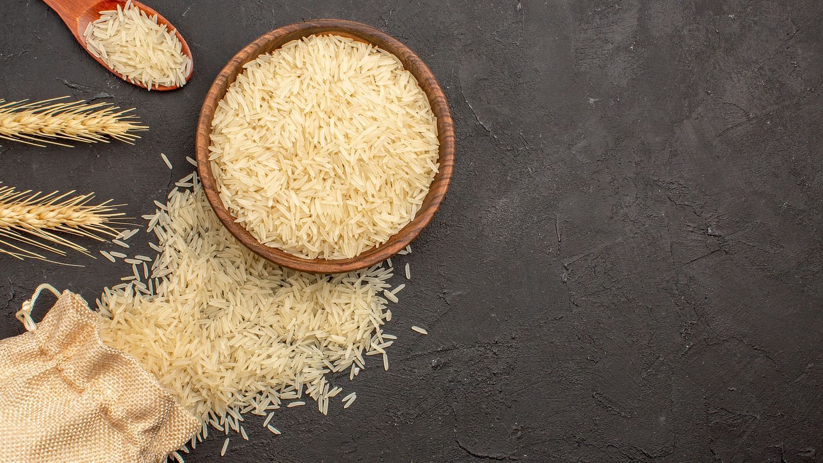 A bowl of uncooked white Basmati rice with a wooden spoon and stalks of paddy on a dark textured background.