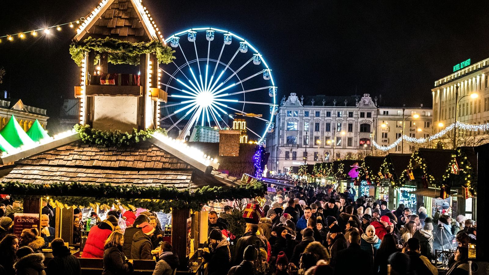 Festive outdoor market at night with a ferris wheel, crowd, and lit stalls, suggesting a holiday fair.