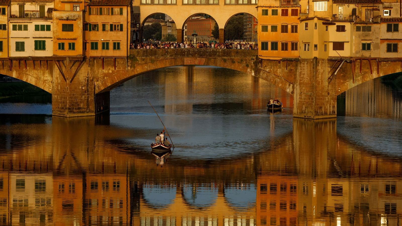 A golden-lit historic stone bridge with arches reflects beautifully on the calm river water during sunset, creating a stunning scenic view.