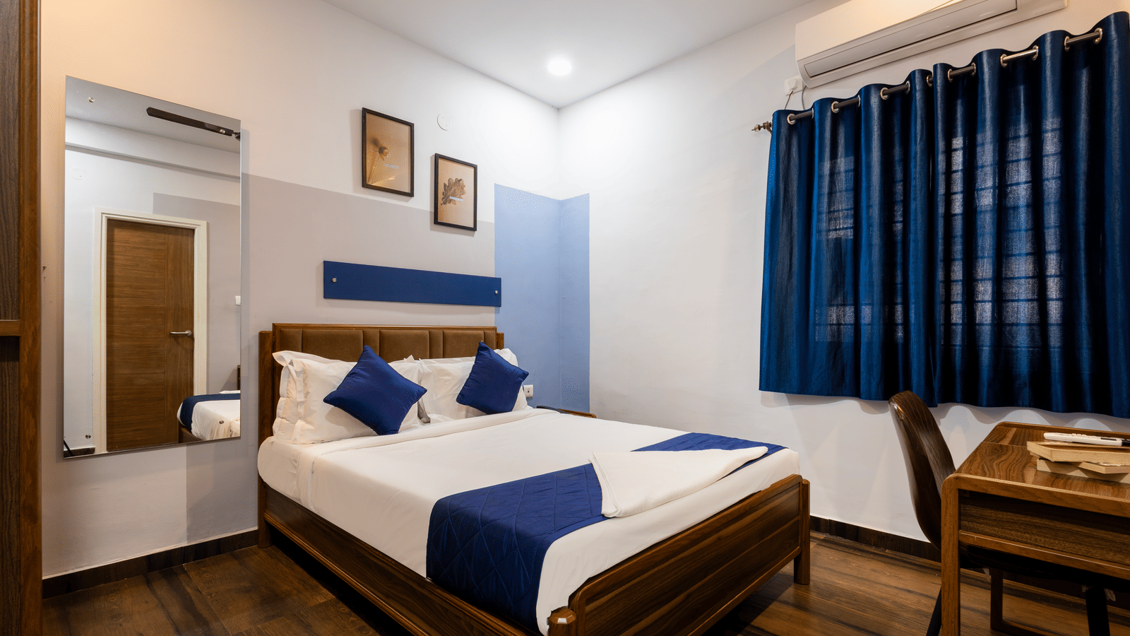 Bedroom in a 1BHK serviced apartment of Kove Smart Stays, seen with wooden flooring, a queen size bed, work desk, and a large wall-mounted mirror.