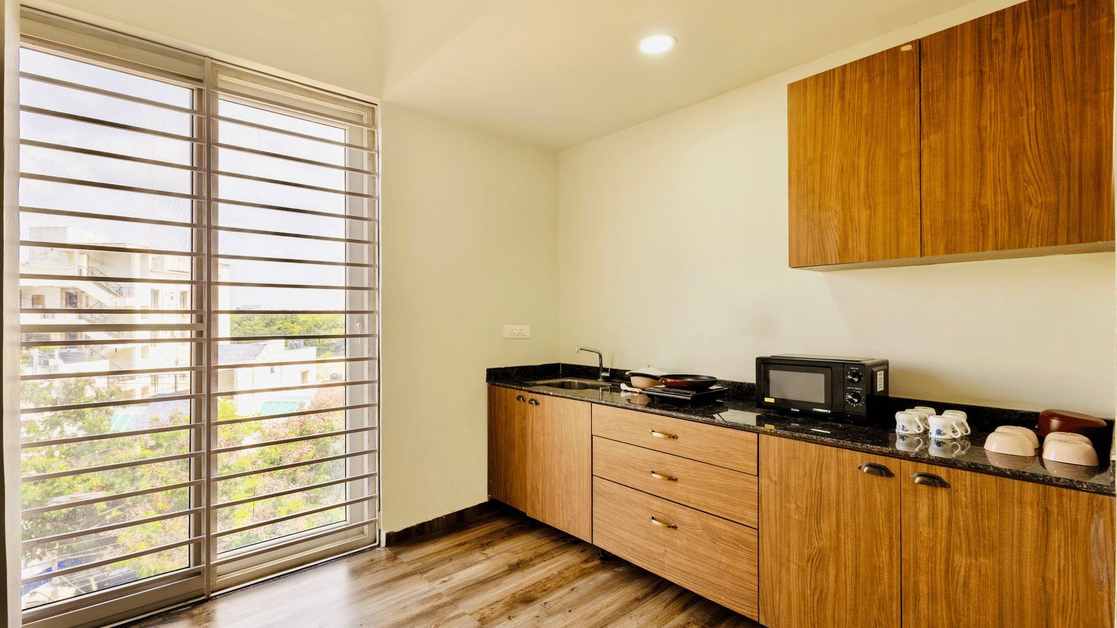 Kitchen set up in one of the 4BHKs at Kove Smart Stays, seen with an oven, wooden flooring, a sink, drawers, wall-mounted shelves, and crockery placed on the countertop.
