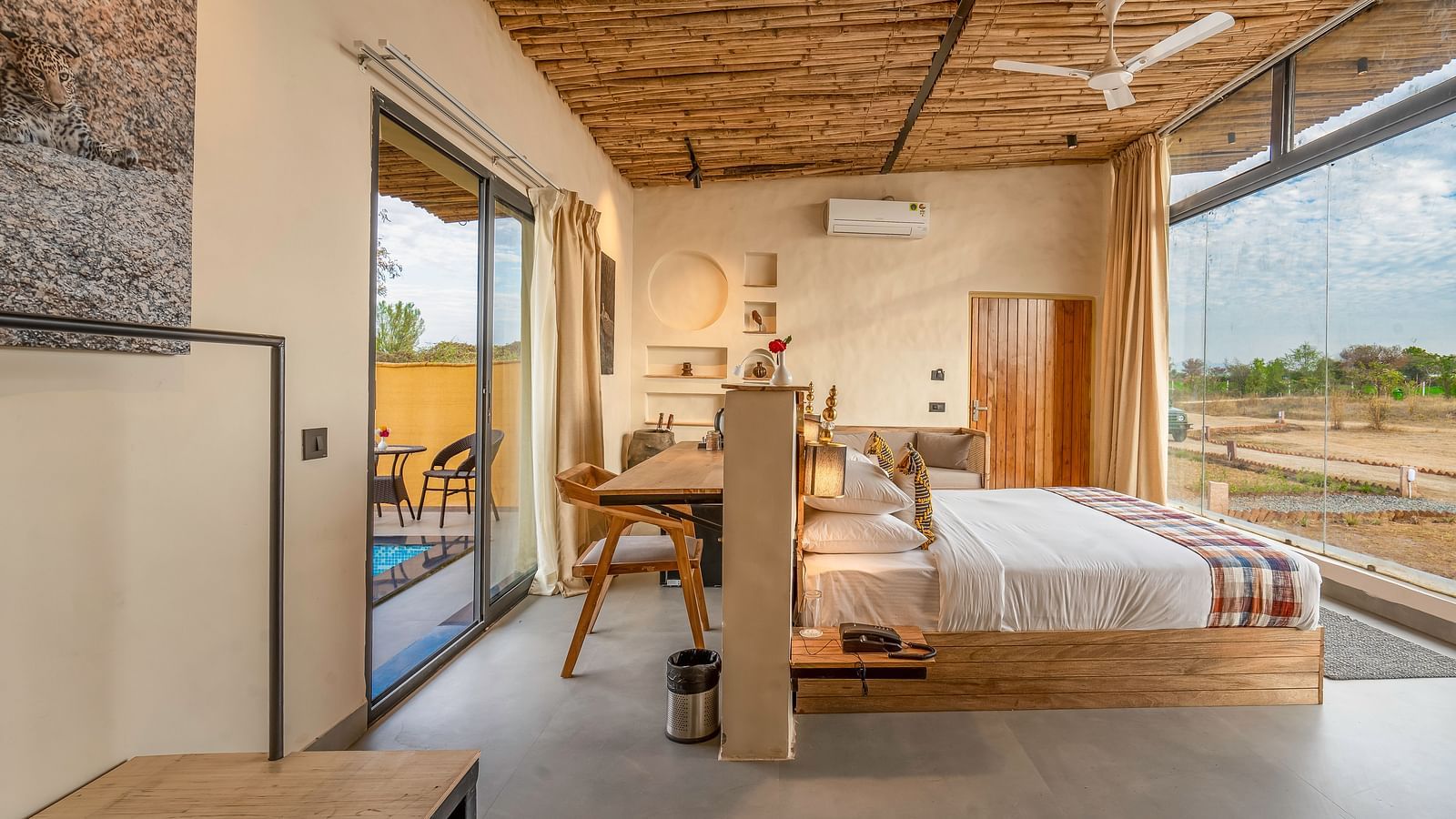 An image of Jungle Cottage bedroom featuring a bed, bedside table with lamp,  a work desk and chair behind, a sofa, AC, ceiling fan, and a large glass door at Kurja Jawai