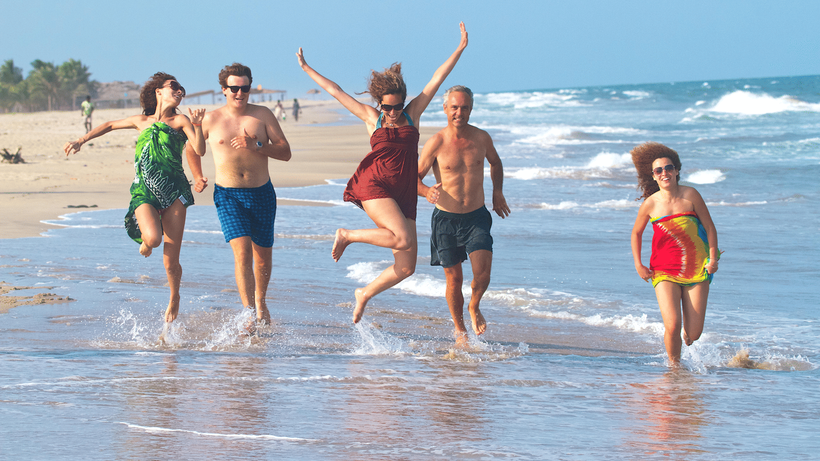 A group of 5 people running and jumping playfully along the shoreline of a sandy beach | Le Pondy