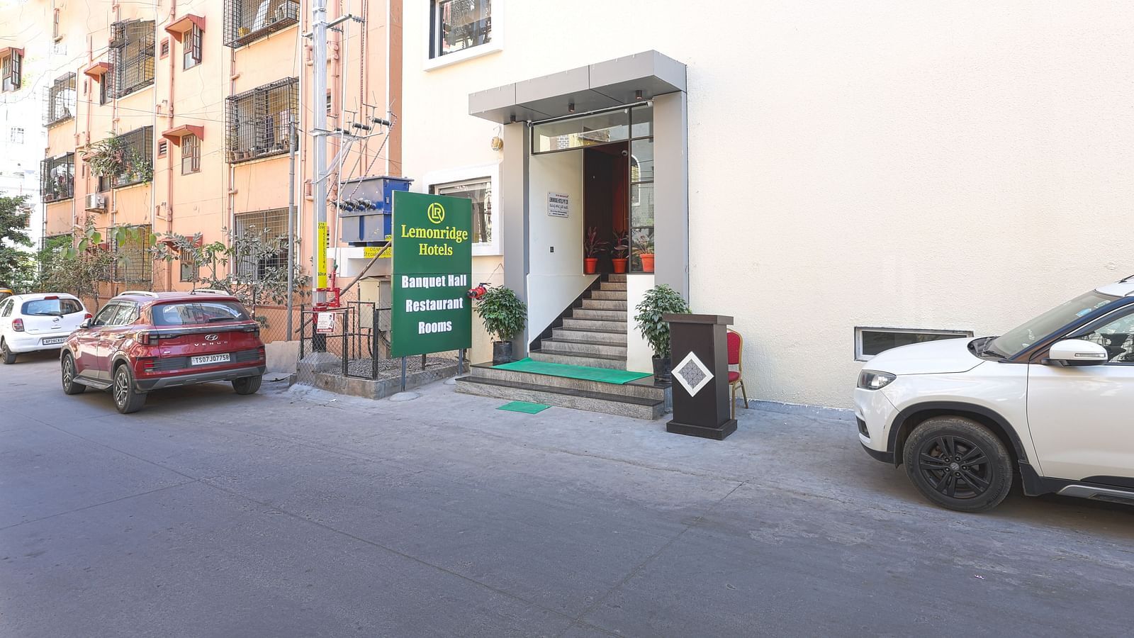 The entrance to the Lemonridge Hotels KPHB property, featuring a signboard that reads 'Lemonridge Hotels' at the top and 'Banquet Halls, Restaurant, Rooms' below. Other buildings are visible on the side, with vehicles parked outside.
