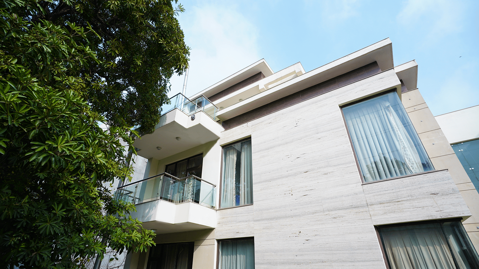 Stone-clad low-rise facade with glass-railed balconies, curtained corner windows and a tree under blue sky.