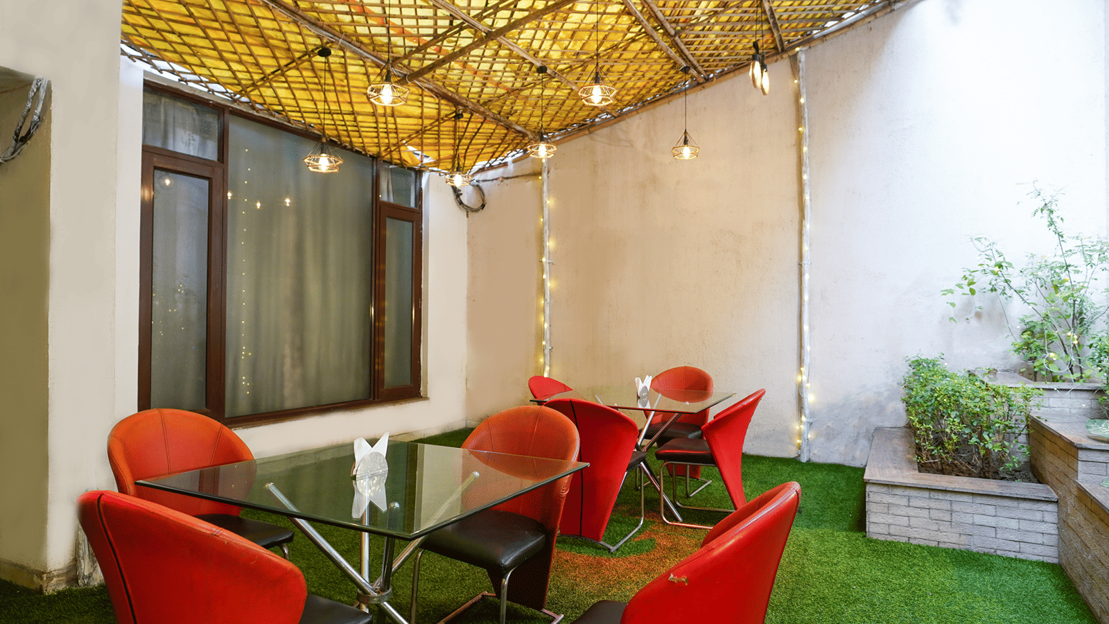 Outdoor courtyard cafe with red tub chairs, glass-top tables, a bamboo-lattice canopy strung with fairy lights, and grass.