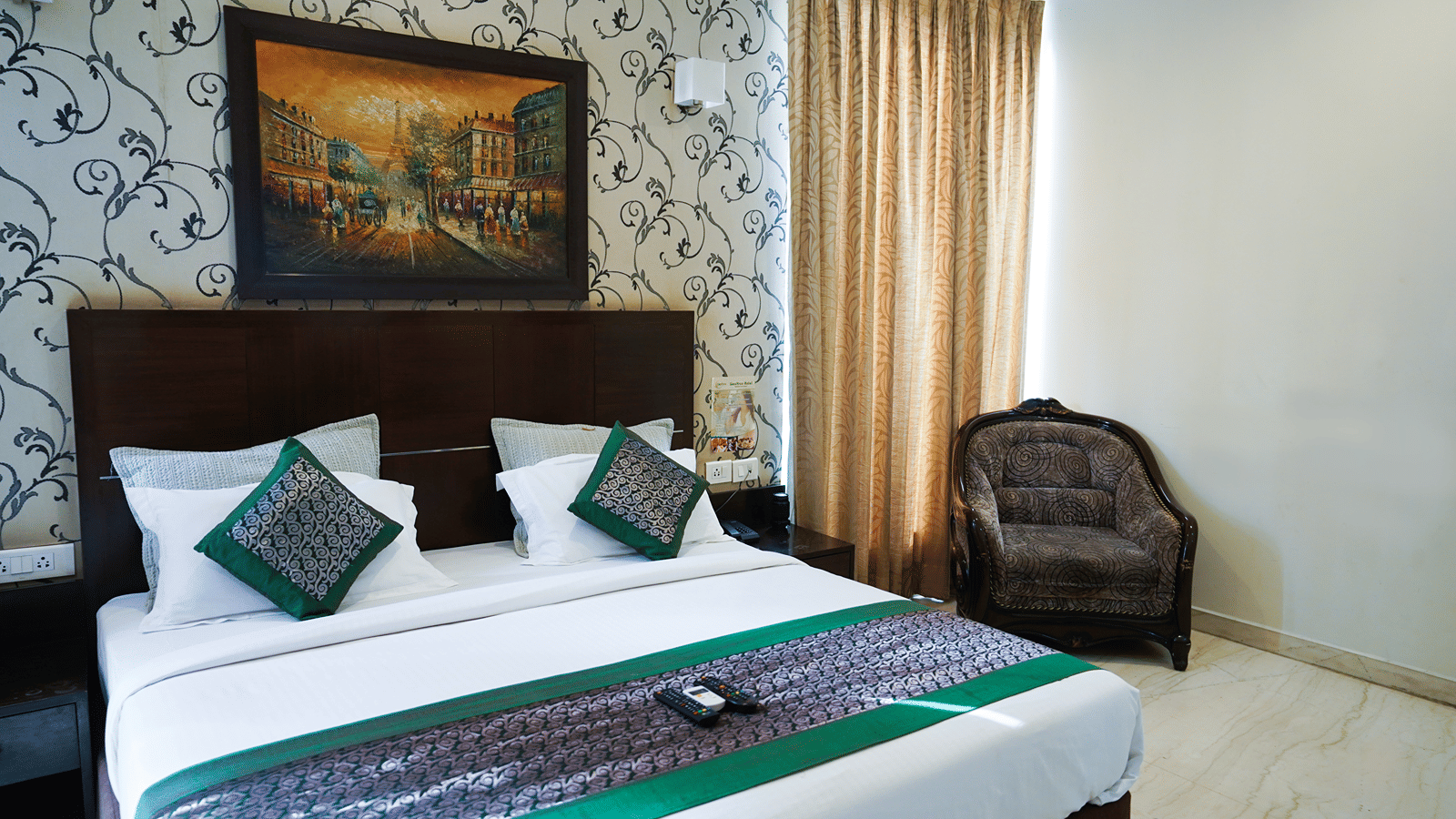 Double bed with crisp linens, elegant patterned walls, a cosy armchair and warm natural light for a relaxing st