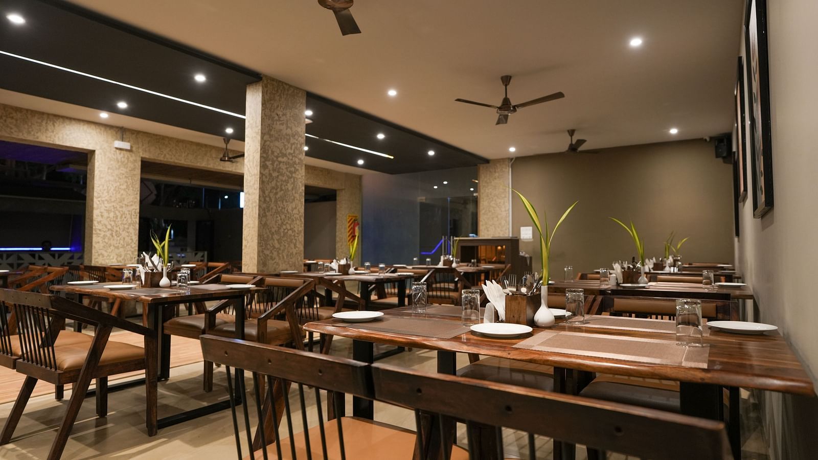 Evening restaurant view with ceiling fans, spotlights, and wooden dining furniture at TGI Livancia Hillside, JC Nagar, Mysore